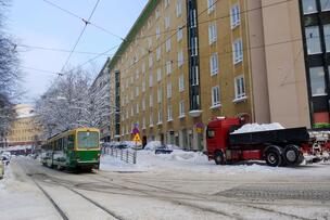 Raitiovaunu seisoo lumisella kadulla. Asuintalojen vieressä on pysäköityjä autoja ja lunta pois vievä kuorma-auto.