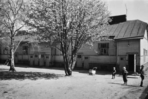 Kuvassa puinen asuintalo sekä pihalla lapsia ja puita.