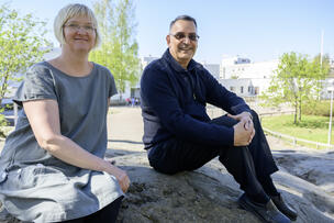 Nainen vasemmalla, mies oikealla katsovat kameraan ja hymyilevät.