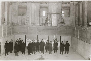 The photo shows 16 men in dark outdoor clothing at the bottom of the unfinished indoor swimming pool. In the background above, a man is plastering a wall.