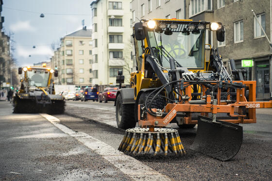 Kaksi työkonetta puhdistaa katua hiekasta, taustalla näkyy kerrostaloja.