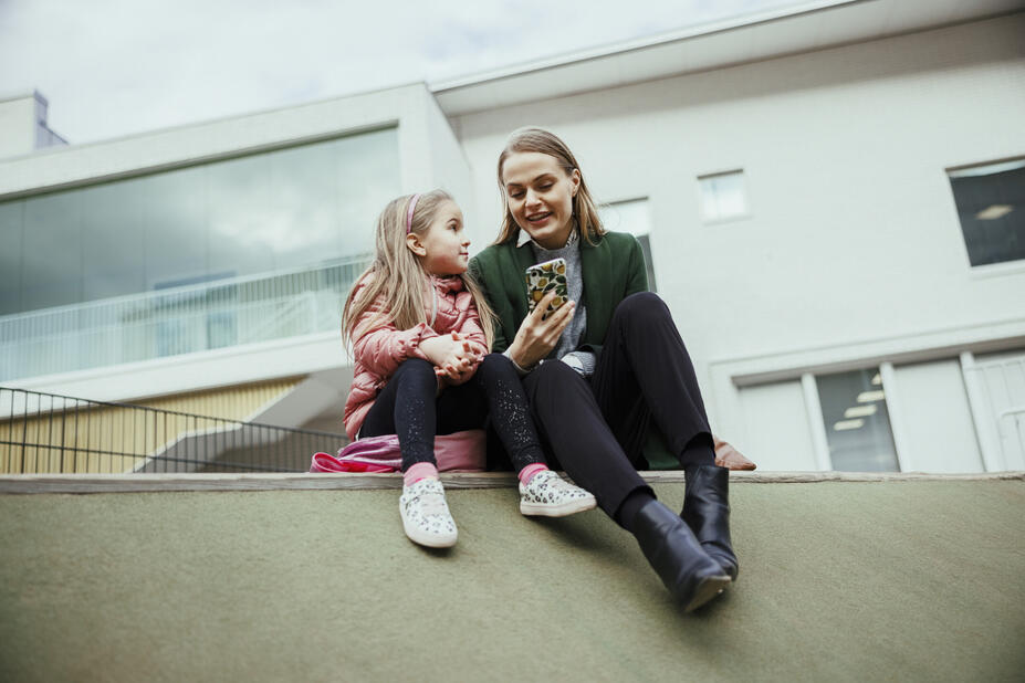 A child and a woman sit outside and look at a mobile device.